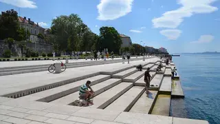 People relaxing on wide stone steps by the seaside with bicycles nearby, under a clear blue sky with scattered clouds.