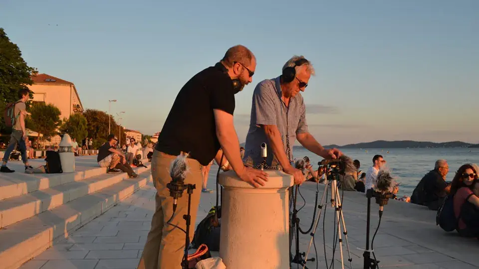 Two men wearing headphones operate audio equipment near a waterfront during sunset, with people sitting on steps nearby.