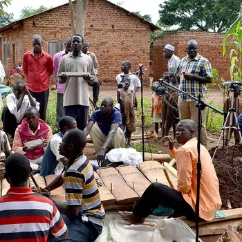 A group of people gathers outdoors around musical instruments and microphones, with a man wearing headphones standing nearby, in a rural setting.